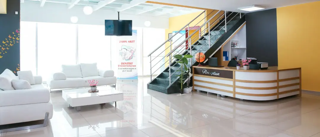 Der Empfangsbereich von Dental Art Alanya mit weißen Sofas, einer Treppe ins Obergeschoss und hohen Fenstern