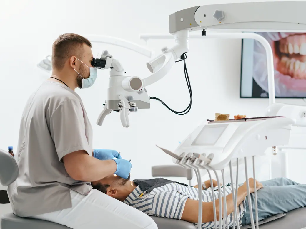 Ein Zahnarzt arbeitet unter einem Dentalmikroskop, dahinter eine intraorale Aufnahme auf dem Bildschirm