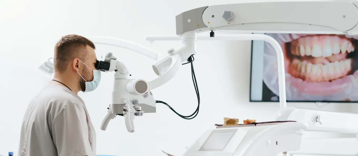 Ein Zahnarzt arbeitet unter einem Dentalmikroskop, dahinter eine intraorale Aufnahme auf dem Bildschirm