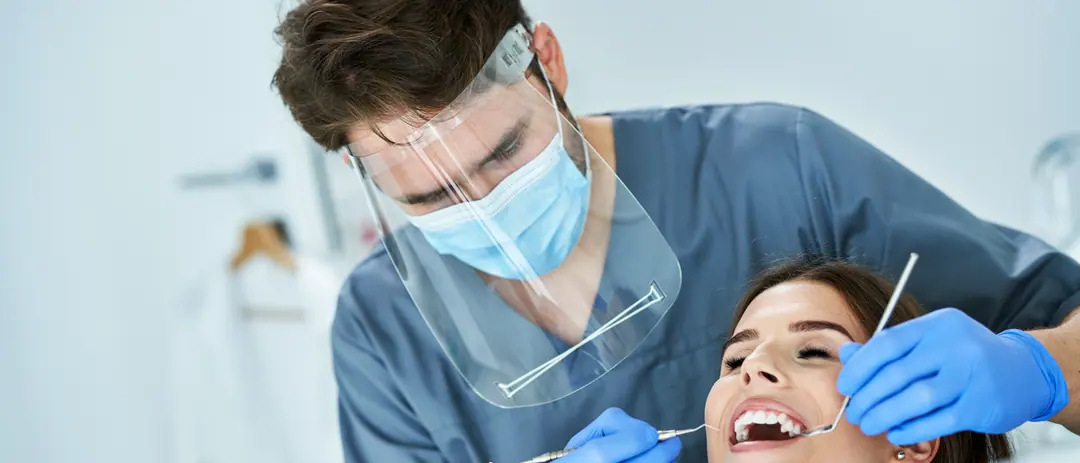 Ein Zahnarzt mit Gesichtsschild untersucht das Zahnfleisch einer lächelnden Patientin